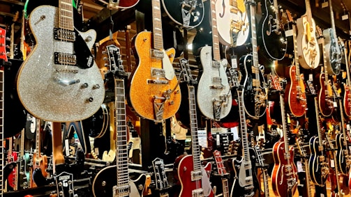 A collection of electric guitars displayed vertically on a wall, showcasing various designs, colors, and finishes. The guitars range from shiny metallic to wooden textures, with intricate details and hardware visible.