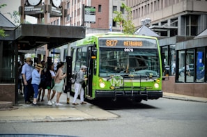 Several people are boarding a bright green city bus at a bus stop in an urban area. The bus displays the route number 807 and the destination 'METROBUS' on its LED sign. The setting features buildings with large glass windows and there is a large clock and signs indicating the place name 'Terminus Place d'Youville' above the bus stop. People appear dressed casually, indicating a relaxed, everyday atmosphere.