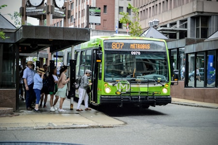 Several people are boarding a bright green city bus at a bus stop in an urban area. The bus displays the route number 807 and the destination 'METROBUS' on its LED sign. The setting features buildings with large glass windows and there is a large clock and signs indicating the place name 'Terminus Place d'Youville' above the bus stop. People appear dressed casually, indicating a relaxed, everyday atmosphere.