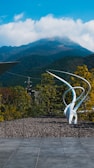 A modern sculpture with elegant, sweeping lines is situated outdoors against a backdrop of lush trees and a distant mountain peak partially covered with clouds. The foreground consists of a tiled surface leading to a stone-paved area.