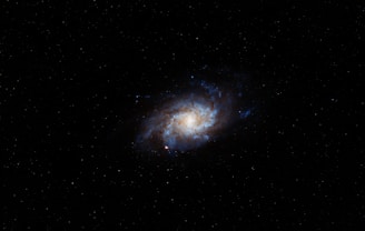 A bright spiral galaxy surrounded by numerous stars scattered across the black void of space. The galaxy has a prominent core with swirling arms radiating outward, exhibiting hues of blue and white.