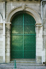 An ornate green metal gate with a circular pattern is framed by an arched stone portal. The surrounding stone wall features classical architectural details, and a small post with a chain is positioned in front.
