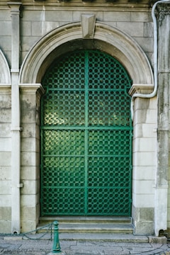 An ornate green metal gate with a circular pattern is framed by an arched stone portal. The surrounding stone wall features classical architectural details, and a small post with a chain is positioned in front.