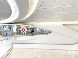 A modern, futuristic interior featuring smooth, flowing architectural lines and a predominantly white color scheme. The space has an abstract design with seamless surfaces and recessed lighting. A large number '1' encased in a red circle is visible in the middle part of the structure. There are glass partitions and metallic details enhancing the sleek aesthetic.