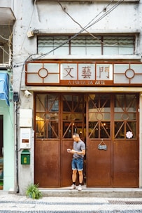 A man stands in front of a brown wooden door of a shop named 'A Porta da Arte'. The building facade is aged with visible wiring and a closed sign is hanging on the door. The man is casually dressed, looking at a phone, and appears relaxed.