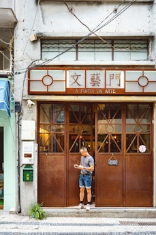 A man stands in front of a brown wooden door of a shop named 'A Porta da Arte'. The building facade is aged with visible wiring and a closed sign is hanging on the door. The man is casually dressed, looking at a phone, and appears relaxed.