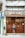 A man stands in front of a brown wooden door of a shop named 'A Porta da Arte'. The building facade is aged with visible wiring and a closed sign is hanging on the door. The man is casually dressed, looking at a phone, and appears relaxed.