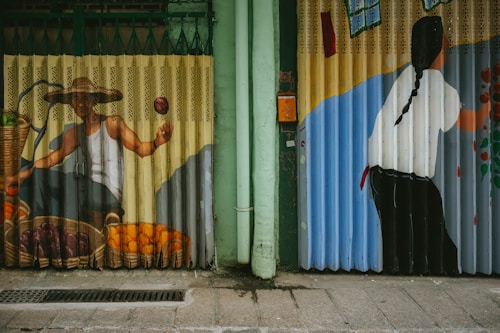 The image features vibrant street art painted on metal shutters. On the left, a figure with a straw hat, wearing a white tank top, holds an apple next to a basket of fruits including oranges and what appear to be plums. On the right, the back of a person with long braided hair is visible, appearing to paint or create art on a blue and beige wall.
