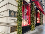 A corner of a luxury retail building with elegant architectural details. The exterior is adorned with festive decorations, including garlands and lights. Large display windows showcase high-end products against a red backdrop. A bright red awning provides shade above the windows, and the street surface appears wet, possibly after rain.