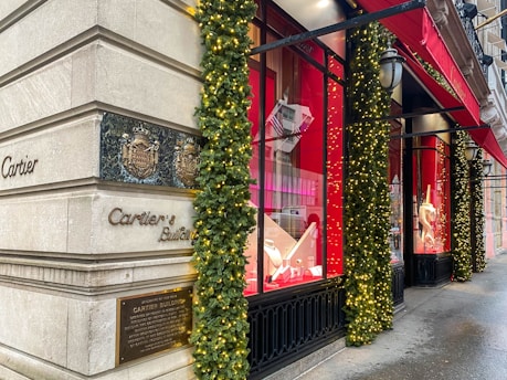 A corner of a luxury retail building with elegant architectural details. The exterior is adorned with festive decorations, including garlands and lights. Large display windows showcase high-end products against a red backdrop. A bright red awning provides shade above the windows, and the street surface appears wet, possibly after rain.