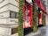 A corner of a luxury retail building with elegant architectural details. The exterior is adorned with festive decorations, including garlands and lights. Large display windows showcase high-end products against a red backdrop. A bright red awning provides shade above the windows, and the street surface appears wet, possibly after rain.