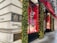 A corner of a luxury retail building with elegant architectural details. The exterior is adorned with festive decorations, including garlands and lights. Large display windows showcase high-end products against a red backdrop. A bright red awning provides shade above the windows, and the street surface appears wet, possibly after rain.