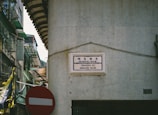 A street sign in Portuguese and Chinese is mounted on a tiled wall at the corner of a building. The area appears to be an urban environment with visible signs of aging infrastructure such as peeling paint and weathered surfaces. A red and white no-entry traffic sign is situated below a tangled cluster of wires. Green awnings and balconies are visible on adjacent buildings, indicating residential or mixed-use structures.
