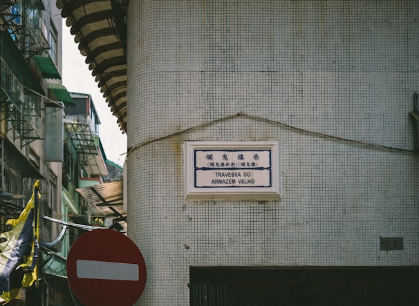A street sign in Portuguese and Chinese is mounted on a tiled wall at the corner of a building. The area appears to be an urban environment with visible signs of aging infrastructure such as peeling paint and weathered surfaces. A red and white no-entry traffic sign is situated below a tangled cluster of wires. Green awnings and balconies are visible on adjacent buildings, indicating residential or mixed-use structures.
