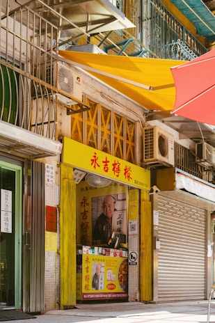 A small shop with a bright yellow and red sign, displaying Chinese characters. The shop has an image of an elderly man on the poster next to offerings of food. There are metal grates and air conditioning units visible above and around the shop. Colorful awnings in red and yellow provide shade.