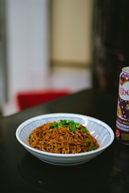 A bowl of crispy fried noodles topped with green onions sits on a dark countertop. Next to it is a can of Ribena, a blackcurrant-flavored beverage. The setting appears to be indoors, perhaps in a kitchen or dining area.