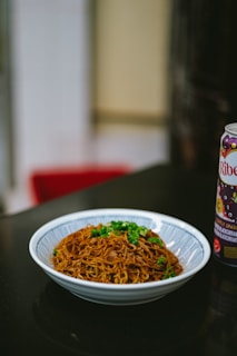 A bowl of crispy fried noodles topped with green onions sits on a dark countertop. Next to it is a can of Ribena, a blackcurrant-flavored beverage. The setting appears to be indoors, perhaps in a kitchen or dining area.