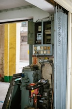 An interior wall features an old electrical panel with several open compartments. Surrounding the panel are multiple electrical sockets and a variety of cords. Below the panel, a red and black industrial drill press is positioned. The area is filled with miscellaneous tools and metal panels, suggesting a functioning workspace. The wall is tiled and adorned with small decorative items, including a strip of red paper with gold lettering.