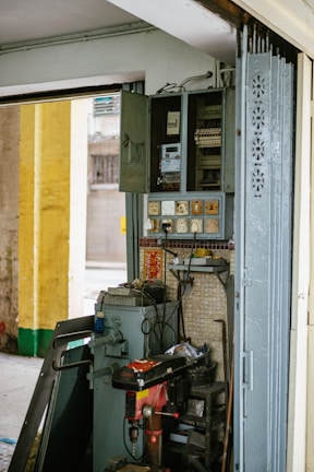 An interior wall features an old electrical panel with several open compartments. Surrounding the panel are multiple electrical sockets and a variety of cords. Below the panel, a red and black industrial drill press is positioned. The area is filled with miscellaneous tools and metal panels, suggesting a functioning workspace. The wall is tiled and adorned with small decorative items, including a strip of red paper with gold lettering.