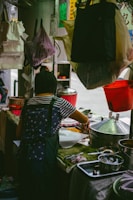 A person wearing a striped shirt and apron is busy preparing food at an outdoor or street food stall. Various kitchen utensils and containers are visible, such as metal pots, trays, and bowls. The setting appears informal with plastic bags and cloths hanging around.