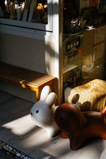 Two cartoon-like animal figures, resembling a rabbit and a dog, are placed outside a store window. The figures are white and brown, respectively, and they are bathed in sunlight. Behind them, various items and colorful posters are visible through the window. A wooden bench and a woven basket add to the cozy and playful setting.