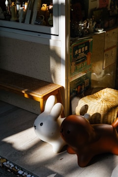 Two cartoon-like animal figures, resembling a rabbit and a dog, are placed outside a store window. The figures are white and brown, respectively, and they are bathed in sunlight. Behind them, various items and colorful posters are visible through the window. A wooden bench and a woven basket add to the cozy and playful setting.