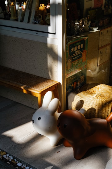 Two cartoon-like animal figures, resembling a rabbit and a dog, are placed outside a store window. The figures are white and brown, respectively, and they are bathed in sunlight. Behind them, various items and colorful posters are visible through the window. A wooden bench and a woven basket add to the cozy and playful setting.