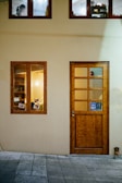 A building facade with a light yellow wall and light wooden door that has a small window. Nearby is a window revealing a warmly lit interior with bookshelves and visible picture frames. Above, two smaller windows show another room with a person wearing a mask working.