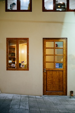 A building facade with a light yellow wall and light wooden door that has a small window. Nearby is a window revealing a warmly lit interior with bookshelves and visible picture frames. Above, two smaller windows show another room with a person wearing a mask working.