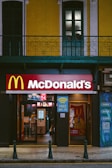 A McDonald's restaurant entrance is visible, situated below a building with bright yellow and white facade and green window frames. Above the entrance, there is a large McDonald's logo. Signs advertising free Wi-Fi and dine-in options are displayed. The entrance showcases glass doors, and there is a glimpse of the interior where menu screens and posters are visible.