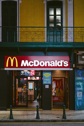 A McDonald's restaurant entrance is visible, situated below a building with bright yellow and white facade and green window frames. Above the entrance, there is a large McDonald's logo. Signs advertising free Wi-Fi and dine-in options are displayed. The entrance showcases glass doors, and there is a glimpse of the interior where menu screens and posters are visible.