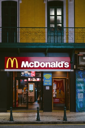A McDonald's restaurant entrance is visible, situated below a building with bright yellow and white facade and green window frames. Above the entrance, there is a large McDonald's logo. Signs advertising free Wi-Fi and dine-in options are displayed. The entrance showcases glass doors, and there is a glimpse of the interior where menu screens and posters are visible.