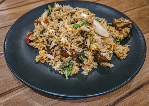 A serving of fried rice is presented on a dark blue plate. The dish includes a mix of vegetables like spinach and chopped green onions, scrambled eggs, and chunks of meat. The rice grains are lightly browned, indicating they have been fried well.