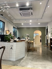 A modern cafe interior with a minimalist design. The front counter is decorated with white tiles and has plants and various items on it. There are ceiling spotlights illuminating the space, and the floor is made of polished concrete. The walls have a light green accent, and there is an archway in the background leading to another room.