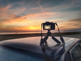 A camera is mounted on a tripod placed on top of a car, capturing a picturesque sunset over a body of water with distant hills. The sky features a blend of warm orange and yellow hues with scattered clouds, creating a serene and captivating atmosphere.