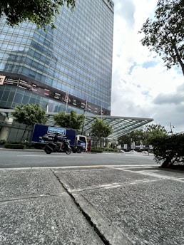 A tall modern glass building with numerous reflective windows stands beside a paved street. In the foreground, a person rides a motorcycle past a delivery truck labeled 'Merrymart Wholesale.' The scene is set under a partly cloudy sky, with trees lining the building and street.