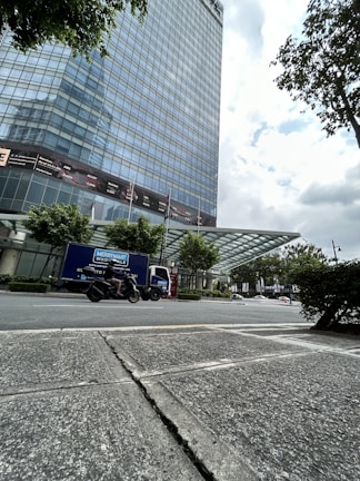 A tall modern glass building with numerous reflective windows stands beside a paved street. In the foreground, a person rides a motorcycle past a delivery truck labeled 'Merrymart Wholesale.' The scene is set under a partly cloudy sky, with trees lining the building and street.