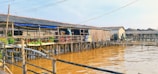 A series of stilt houses built over a body of murky brown water, connected by wooden walkways. The structures are constructed with wooden beams and tin roofs, and some have hanging clothes and potted plants for decoration. The setting appears to be in a rustic, possibly tropical location.