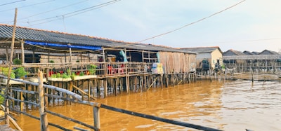 A series of stilt houses built over a body of murky brown water, connected by wooden walkways. The structures are constructed with wooden beams and tin roofs, and some have hanging clothes and potted plants for decoration. The setting appears to be in a rustic, possibly tropical location.