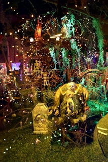 A spooky, dimly lit Halloween scene with various decorations including skeletons and tombstones. Strings of colorful lights are draped across the setup, casting eerie glows. A large skeleton figure sits prominently in the foreground with a sign that reads 'Chill, the chill don't disturb.' The background is filled with more lights and Halloween-themed figures, creating a densely packed, festive atmosphere.