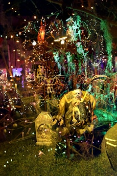 A spooky, dimly lit Halloween scene with various decorations including skeletons and tombstones. Strings of colorful lights are draped across the setup, casting eerie glows. A large skeleton figure sits prominently in the foreground with a sign that reads 'Chill, the chill don't disturb.' The background is filled with more lights and Halloween-themed figures, creating a densely packed, festive atmosphere.