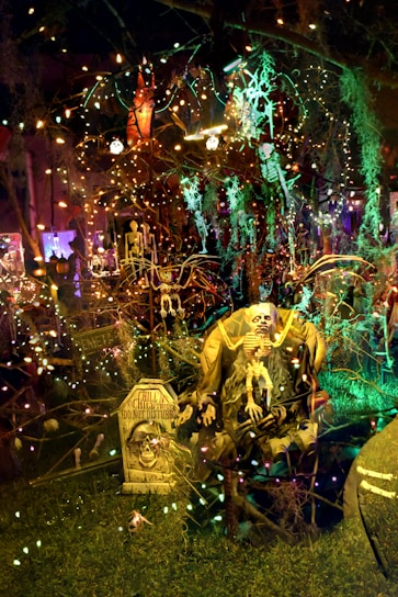 A spooky, dimly lit Halloween scene with various decorations including skeletons and tombstones. Strings of colorful lights are draped across the setup, casting eerie glows. A large skeleton figure sits prominently in the foreground with a sign that reads 'Chill, the chill don't disturb.' The background is filled with more lights and Halloween-themed figures, creating a densely packed, festive atmosphere.