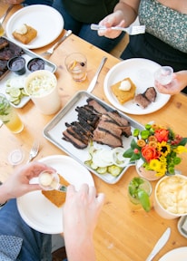 A wooden table is set with plates of food, including slices of brisket, pickles, and cornbread with butter. Drinks are served in glasses, and a vibrant bouquet of flowers adds a pop of color. People are serving food onto their plates with utensils.