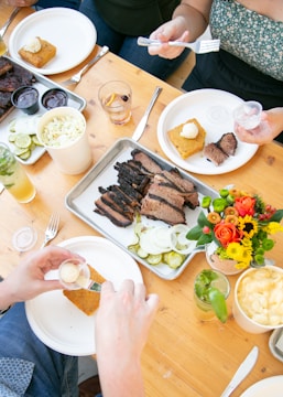 A wooden table is set with plates of food, including slices of brisket, pickles, and cornbread with butter. Drinks are served in glasses, and a vibrant bouquet of flowers adds a pop of color. People are serving food onto their plates with utensils.