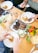 A wooden table is set with plates of food, including slices of brisket, pickles, and cornbread with butter. Drinks are served in glasses, and a vibrant bouquet of flowers adds a pop of color. People are serving food onto their plates with utensils.