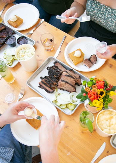 A wooden table is set with plates of food, including slices of brisket, pickles, and cornbread with butter. Drinks are served in glasses, and a vibrant bouquet of flowers adds a pop of color. People are serving food onto their plates with utensils.