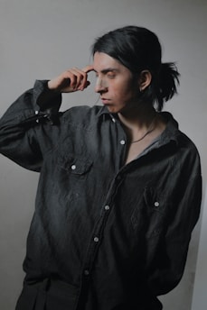 A person with long dark hair tied back, wearing a dark denim shirt with button closures, and a contemplative expression. A patch is visible on the nose, suggesting recent injury or healing. The background is a plain, neutral tone, enhancing the focus on the subject.