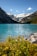 A serene mountain landscape featuring a turquoise lake surrounded by high snow-capped peaks and lush green forests. In the foreground, bright yellow wildflowers and rocky terrain add color and texture.