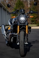 A close-up view of a modern motorcycle with a round front headlight and sleek design. The motorcycle features golden front forks, black handlebars, and a robust exhaust system. The background includes a blurred view of trees and part of a building, emphasizing the motorcycle as the main subject.