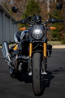 A close-up view of a modern motorcycle with a round front headlight and sleek design. The motorcycle features golden front forks, black handlebars, and a robust exhaust system. The background includes a blurred view of trees and part of a building, emphasizing the motorcycle as the main subject.
